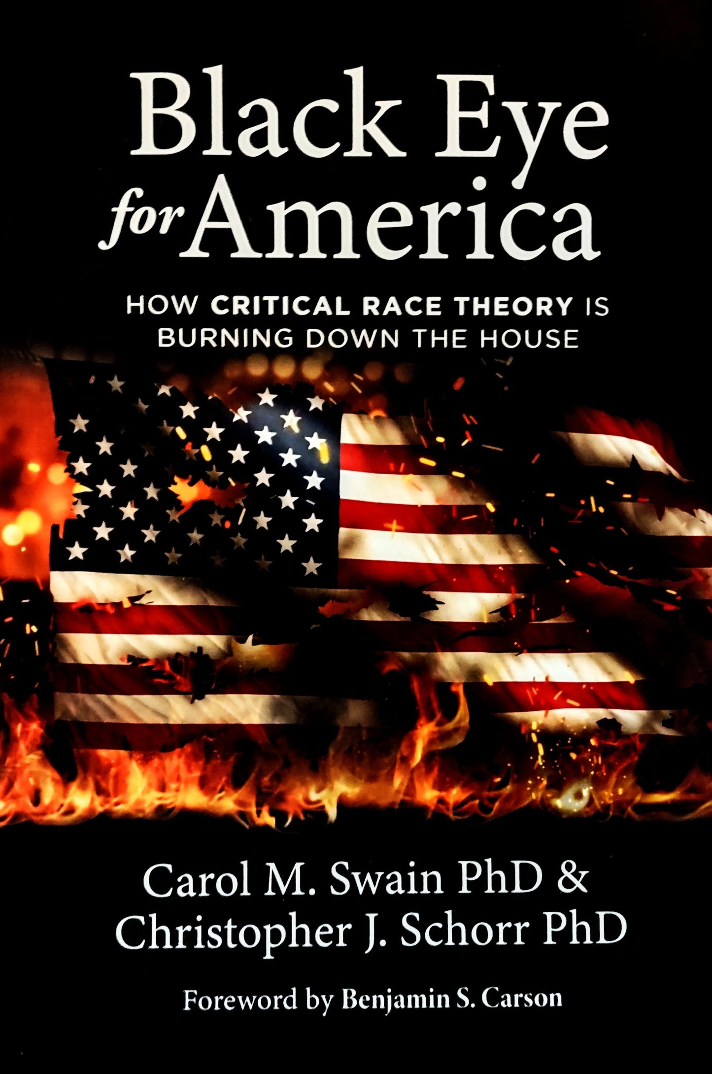 Book cover of 'Black Eye for America: How Critical Race Theory is Burning Down the House (ISBN: 9781737419808)' by Carol Swain and Christopher Schorr and foreword by Ben Carson, with a burning American flag on a black background.