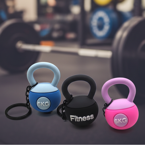 Three small kettlebell-shaped fitness accessories in light blue, black, and pink are shown on a gym floor with a blurred barbell in the background. Each has a keychain ring and a circular badge reading 6KG; the center item also has the word Fitness on it.