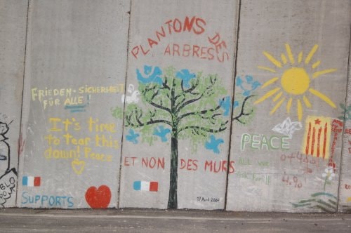 Photograph of political graffiti on the West Bank Wall in Israel/Palestine, featuring text and symbols related to peace and resistance.