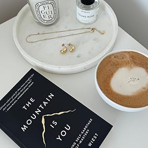 Book 'The Mountain is You' by Brianna Wiest with a cup of coffee and a necklace on a table.