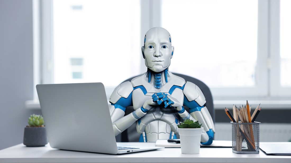 Humanoid robot sitting at a desk in front of a laptop appearing engaged and interested.