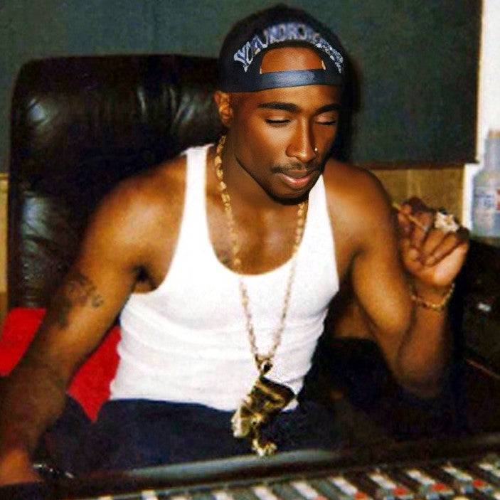 Tupac Shakur in sitting in a chair in front of the mixing board in the studio smoking, wearing a backwards snapback and tank top as well as a gold chain with a Nefertiti pendant.