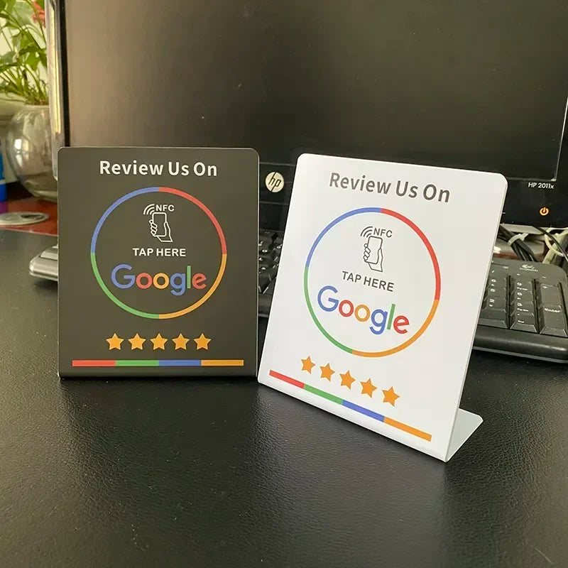 Two 'Review Us On Google' signboards on a desk with a computer in the background.