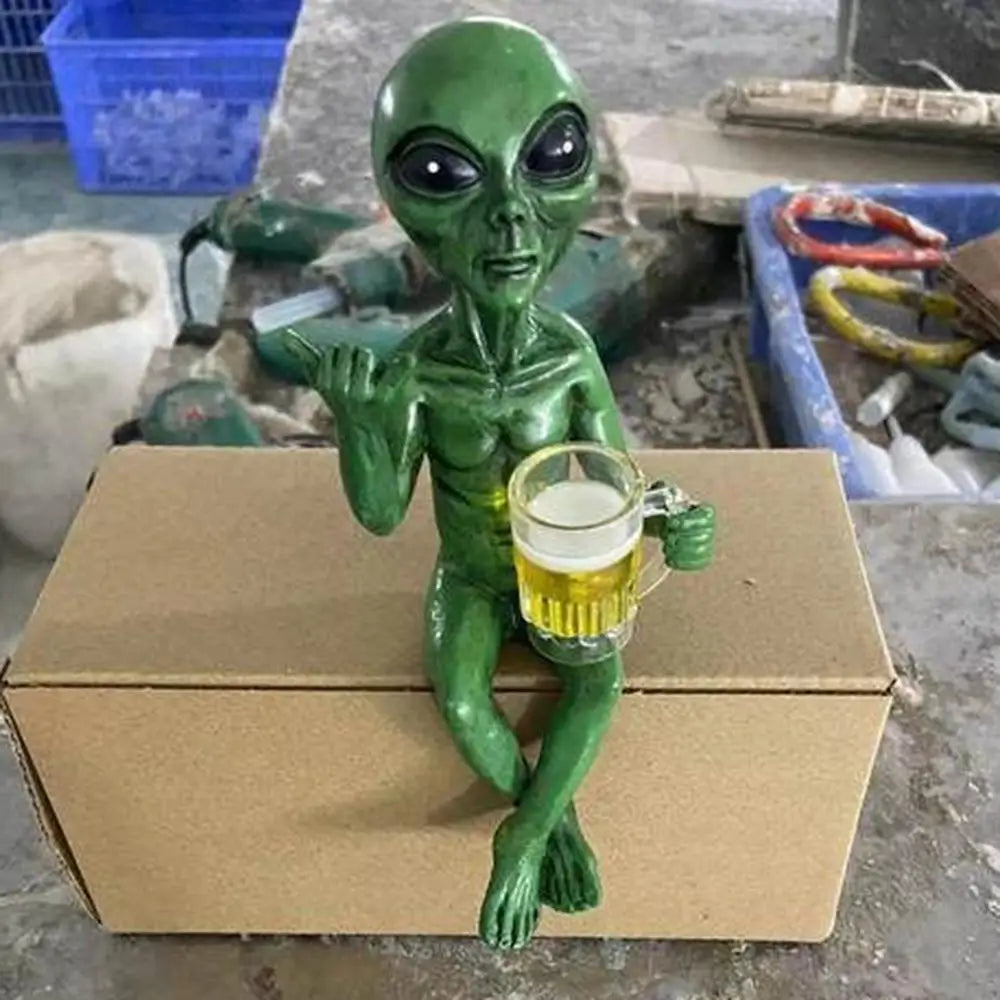 Green alien figurine holding a glass of beer on a cardboard box