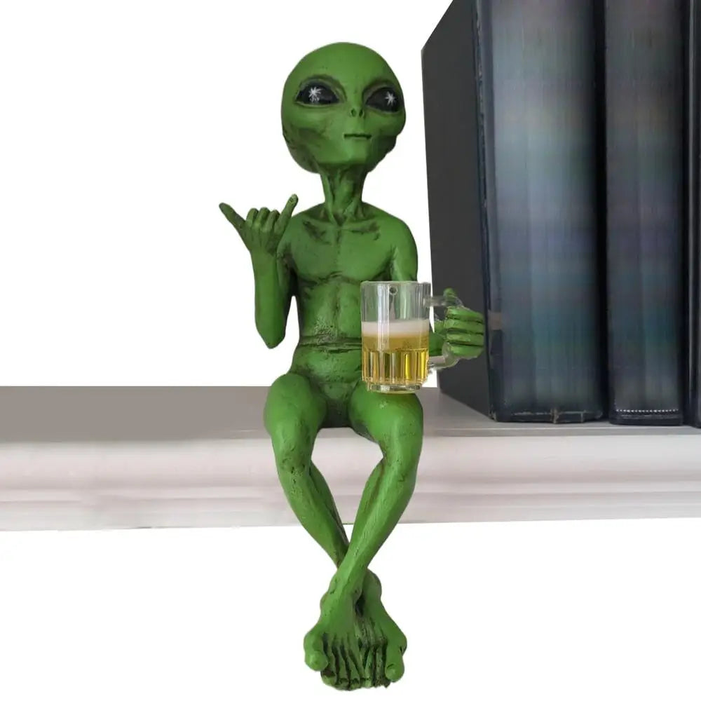Laid-back green alien figurine sitting cross-legged on a bookshelf, holding a beer mug and making the shaka sign.