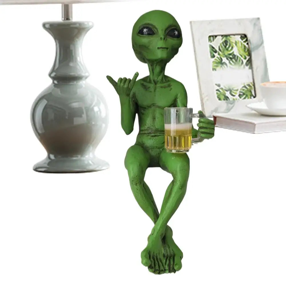 Laid-back green alien figurine sitting cross-legged next to other decor, holding a beer mug and making the shaka sign.
