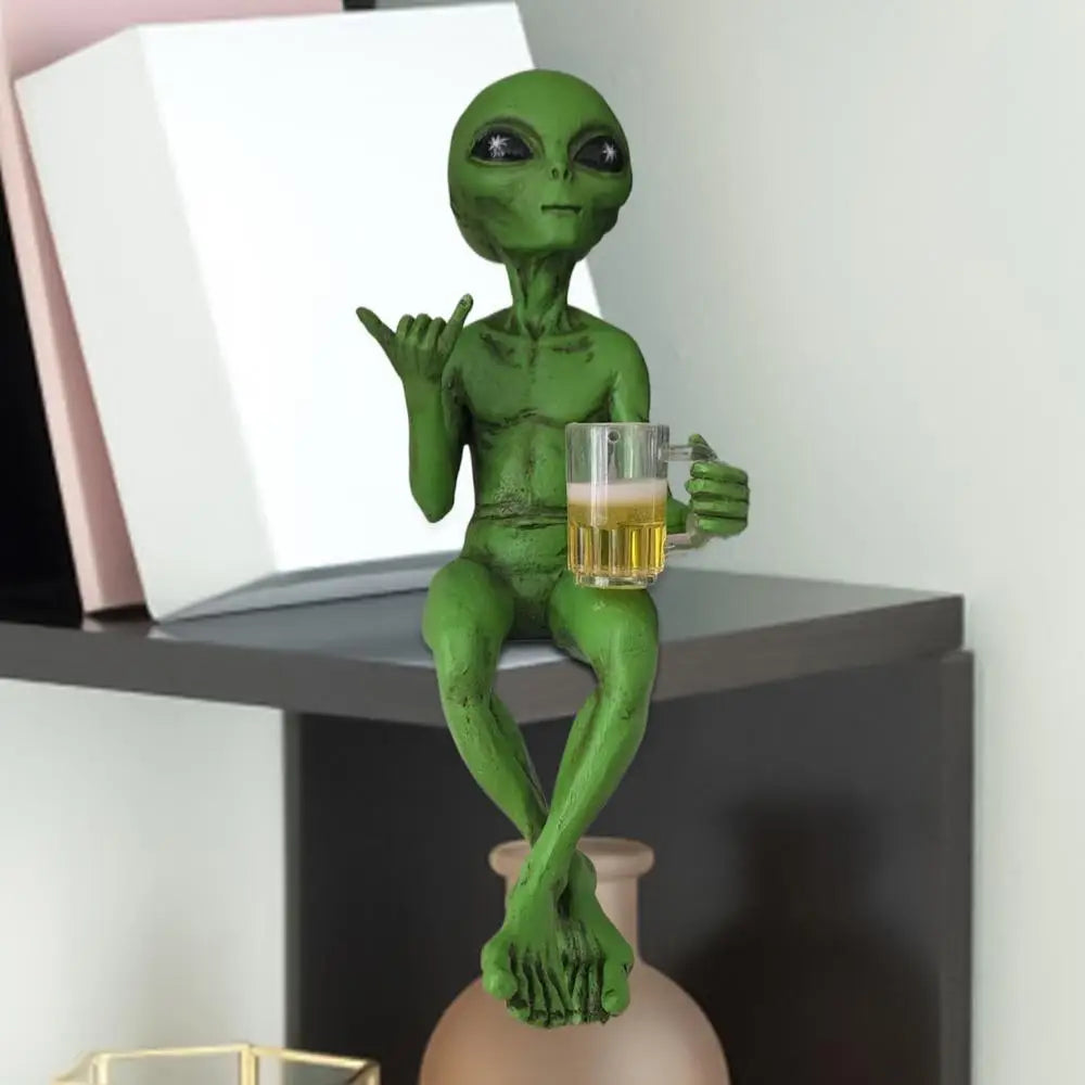Laid-back green alien figurine sitting cross-legged on a bookshelf corner, holding a beer mug and making the shaka sign.