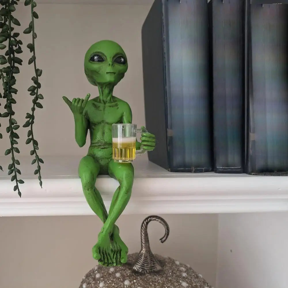Laid-back green alien figurine sitting cross-legged on a bookshelf, holding a beer mug and making the shaka sign.