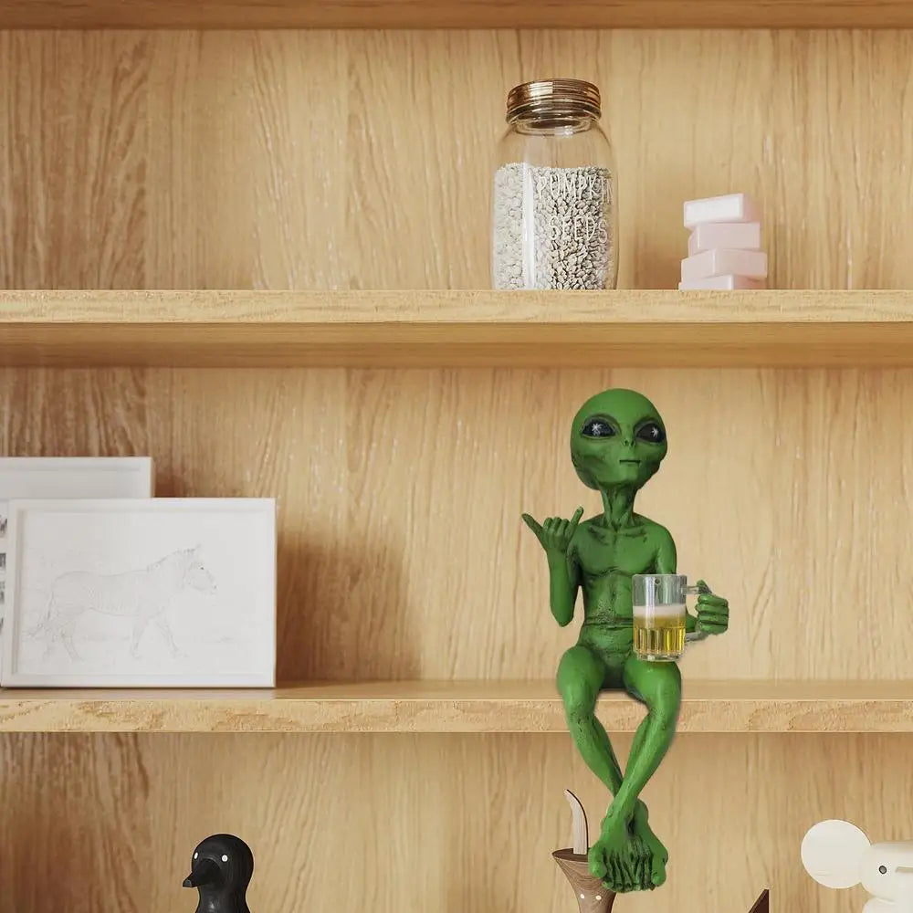 Laid-back green alien figurine sitting cross-legged on a bookshelf, holding a beer mug and making the shaka sign.