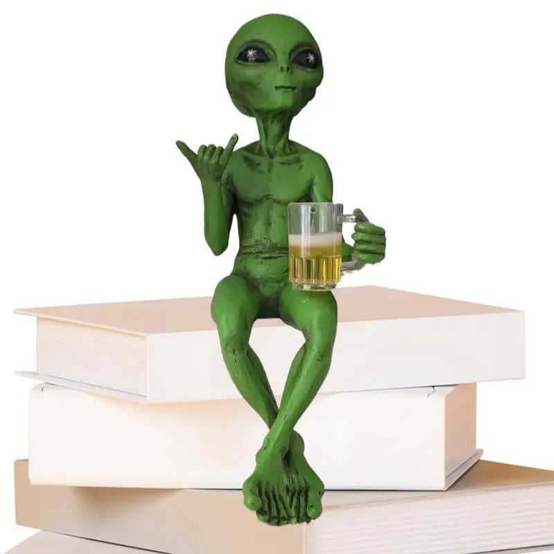 Green alien figurine holding a mug of beer with his left hand and the shaka "hang-10" sign with his right hand sitting on a stack of books with a white background