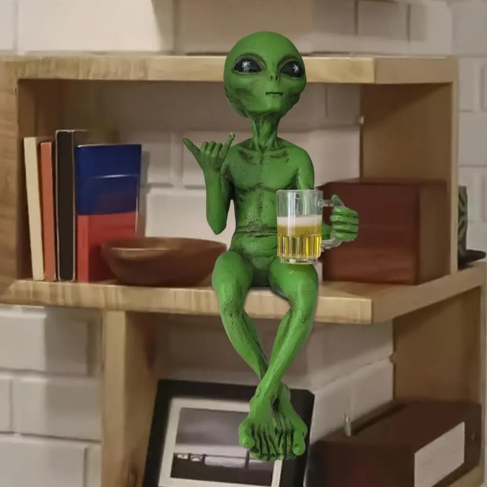 Laid-back green alien figurine sitting cross-legged on a bookshelf corner, holding a beer mug and making the shaka sign.