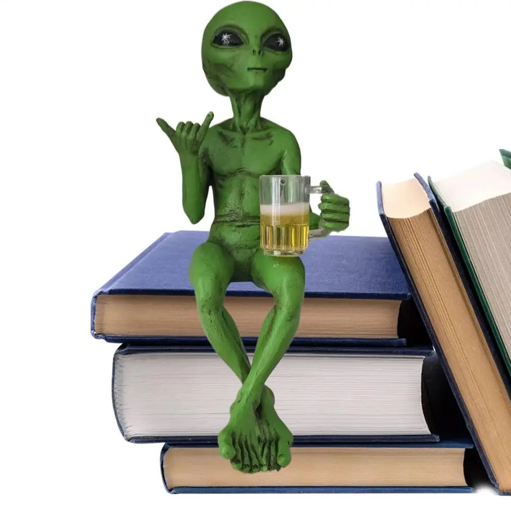Laid-back green alien figurine sitting cross-legged on a stack of blue books, holding a beer mug and making the shaka sign.