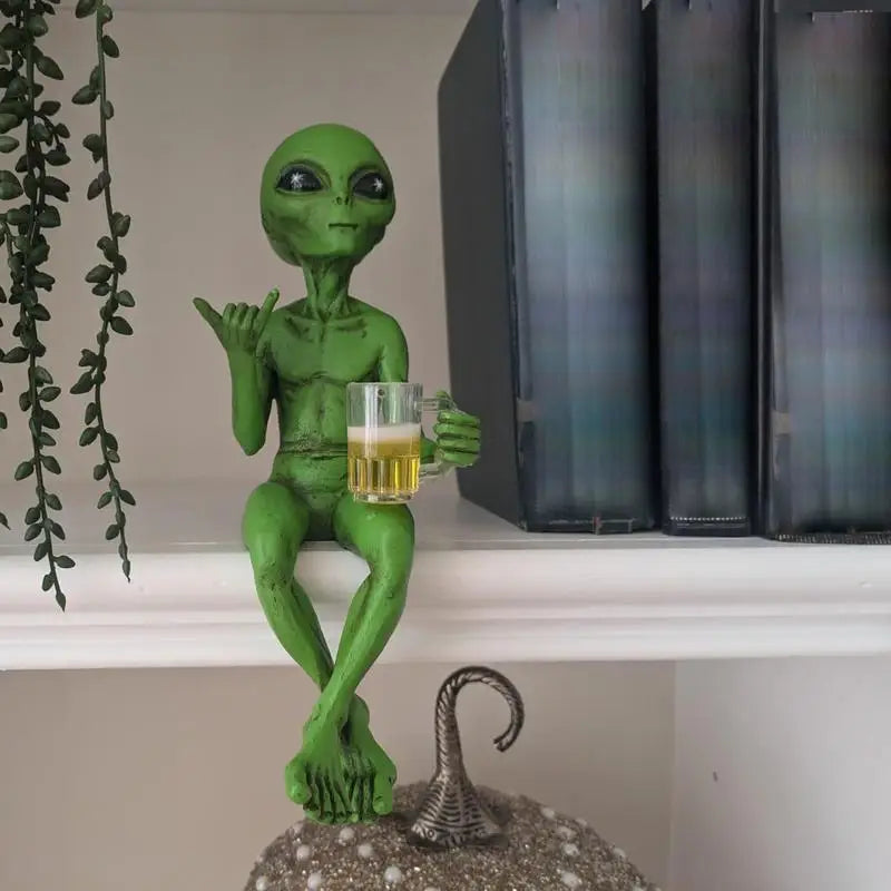 Laid-back green alien figurine sitting cross-legged on a bookshelf, holding a beer mug and making the shaka sign.