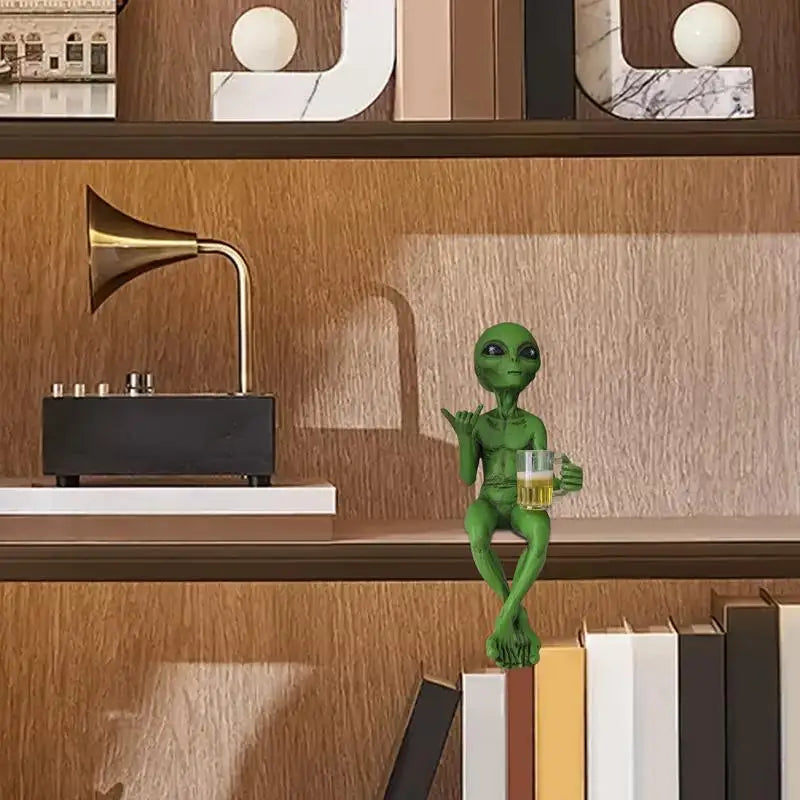 Laid-back green alien figurine sitting cross-legged on a bookshelf, holding a beer mug and making the shaka sign.