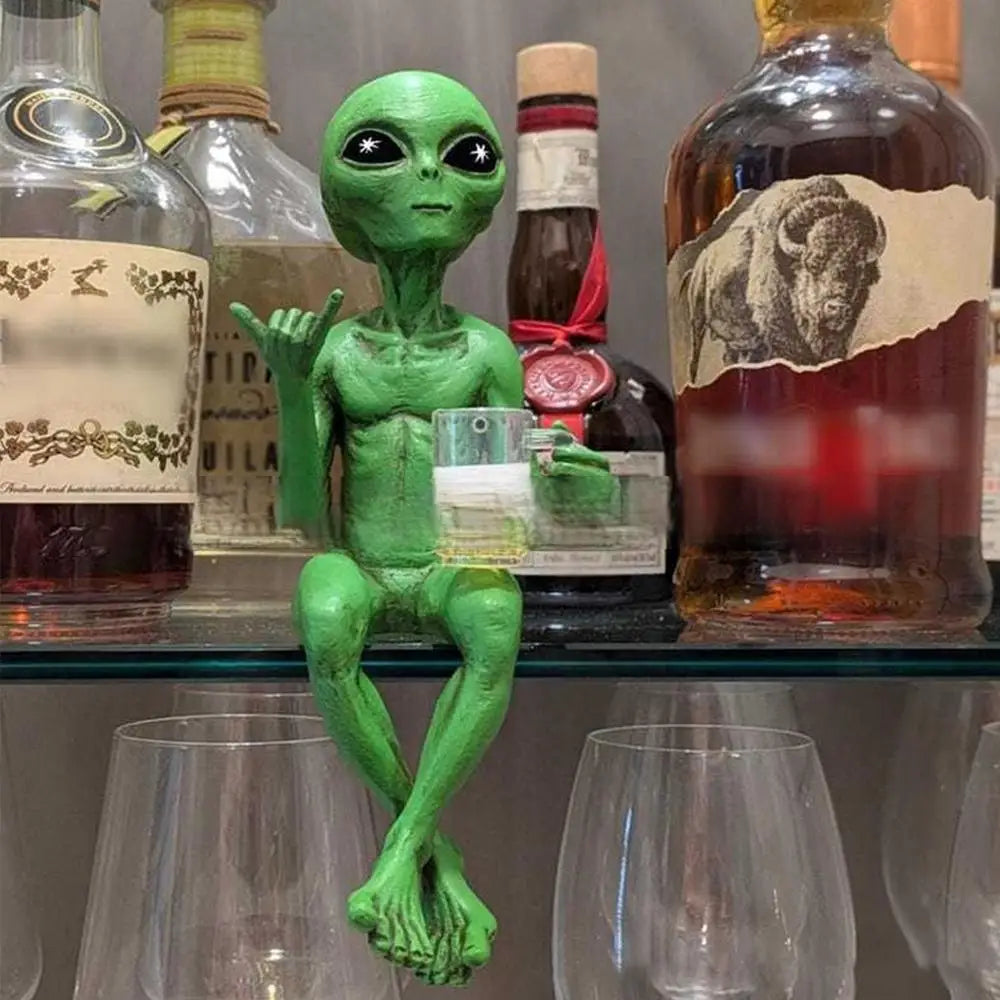 Laid-back green alien figurine sitting cross-legged on a liquor cabinet shelf, holding a beer mug and making the shaka sign.