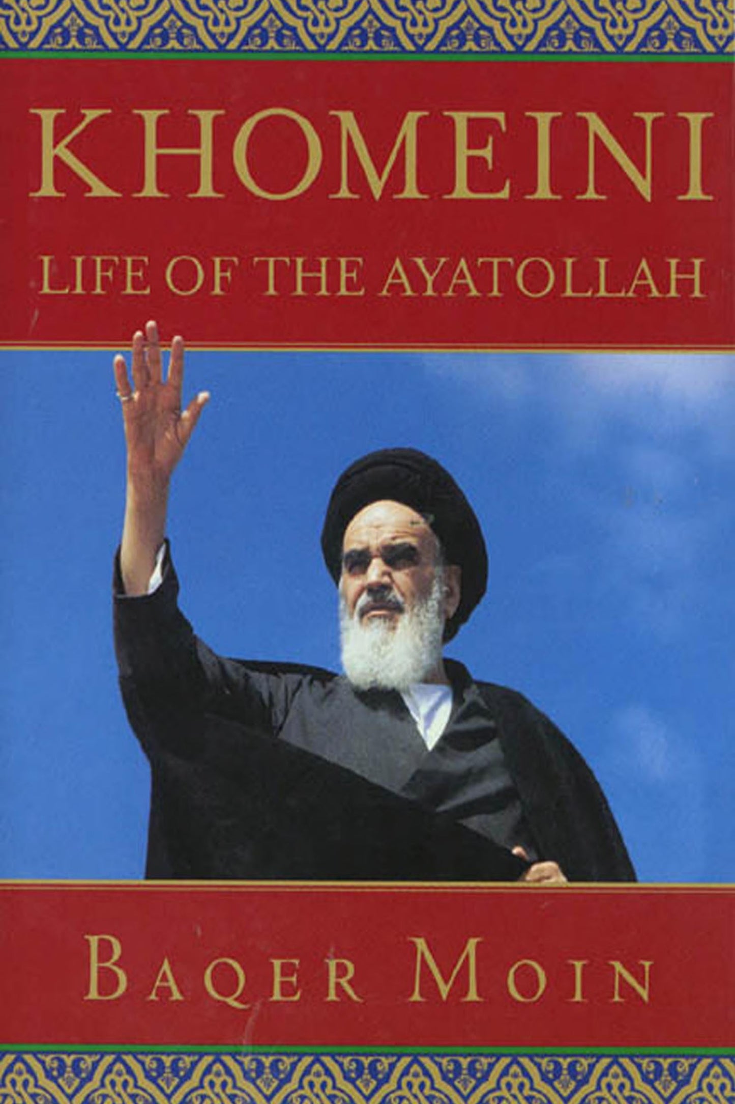 Book cover for 'Khomeini: Life of the Ayatollah' by Baqer Moin, featuring a photo of Ayatollah Khomeini raising his hand against a blue sky, framed by red title bands and ornate gold-and-blue patterned borders.