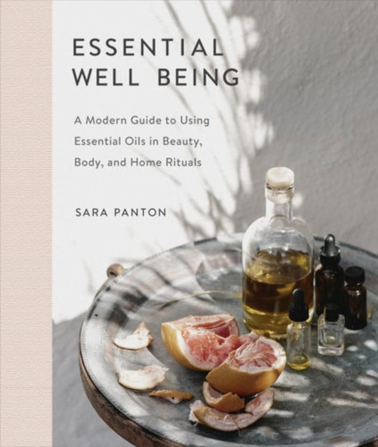 Book cover of 'Essential Well Being: A Modern Guide to Using Essential Oils in Beauty, Body, and Home Rituals' by Sara Panton with essential oils and fruit on a table.