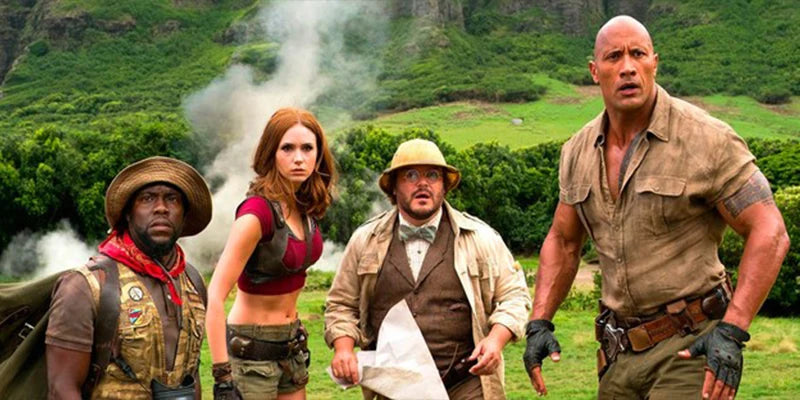 Four characters from jumanji movie standing in a lush green outdoor setting with mountains in the background.