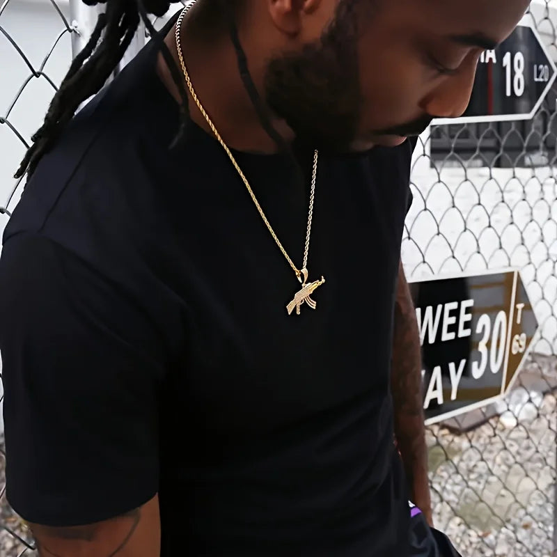 Man wearing a black shirt and gold necklace with AK47 rifle pendant, standing in front of a chain-link fence.