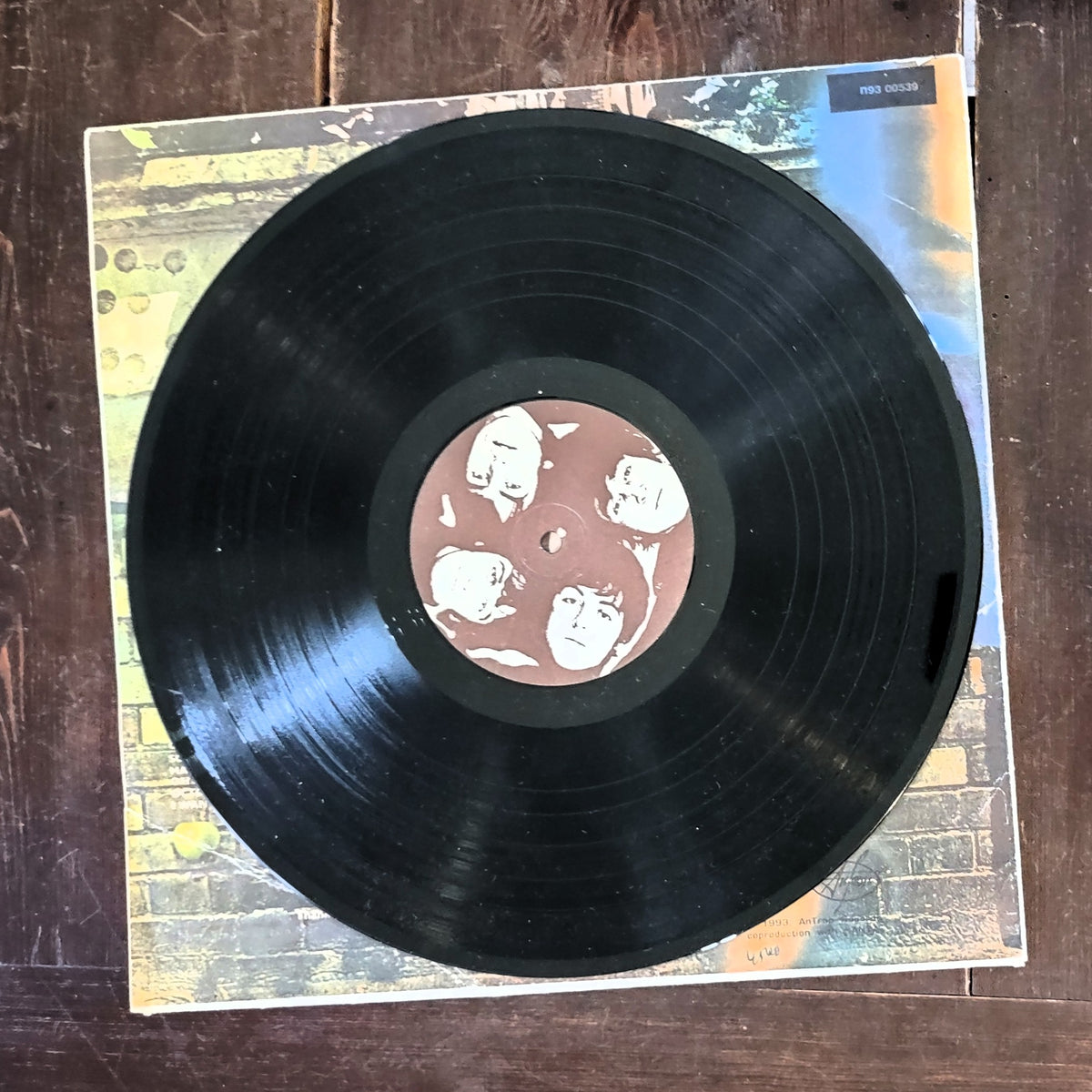 Black Beatles Abbey Road vinyl record partially pulled from its illustrated album cover, placed on a dark wooden surface. The center label shows the faces of four children, and the cover art has a colorful abstract background.