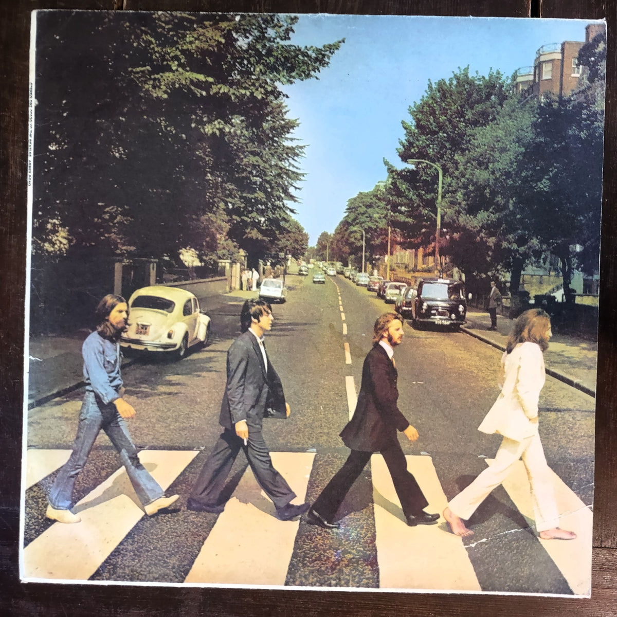 The Beatles' Abbey Road album cover shown flat against a dark background, featuring the four band members walking across a zebra road crossing on a tree-lined street with classic cars.
