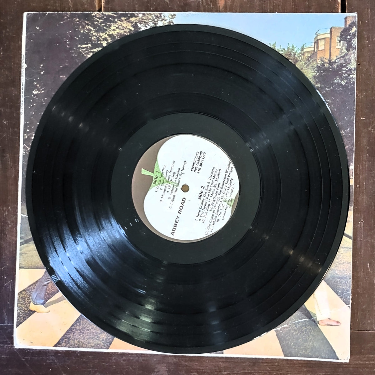 A black vinyl record partially pulled from its printed album cover, photographed from above on a dark wooden surface. The center label reads 'Abbey Road (Side-1, Side-2) Beatles.