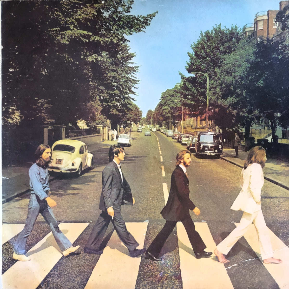 The Abbey Road - Beatles album cover artwork featuring the four band members walking across a zebra crossing on a tree-lined street, with vintage cars and a blue sky in the background.