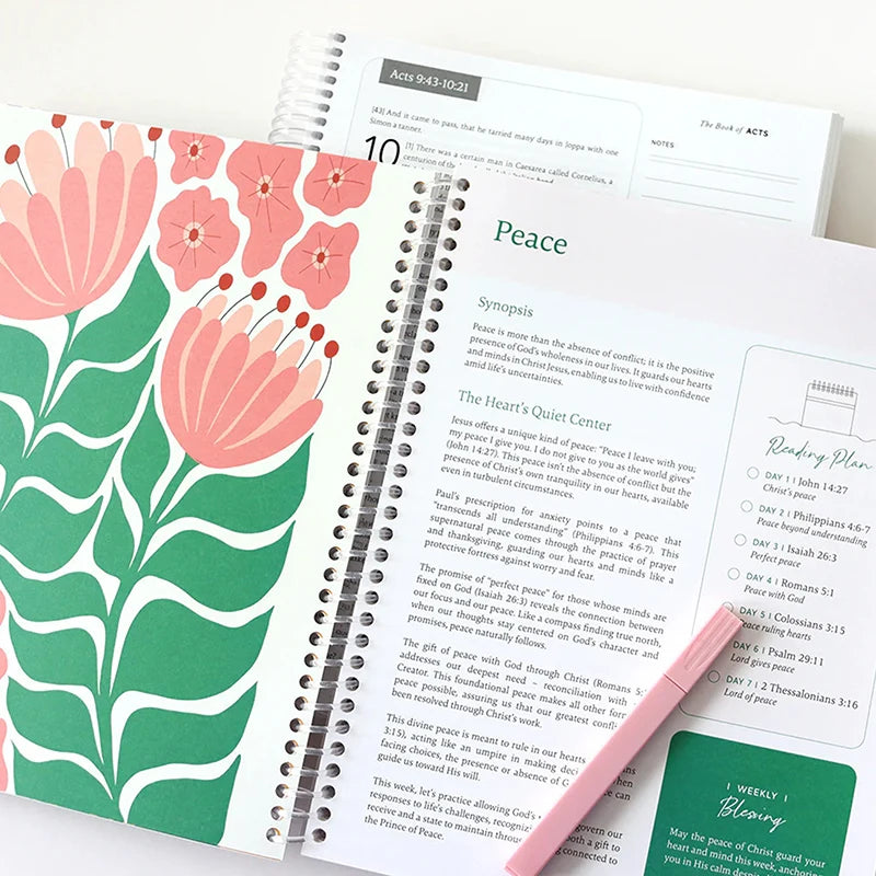 Open planner book 'The Bible in a Year' by Spiral Bible with floral design on left page and text on right page, pink pen on top