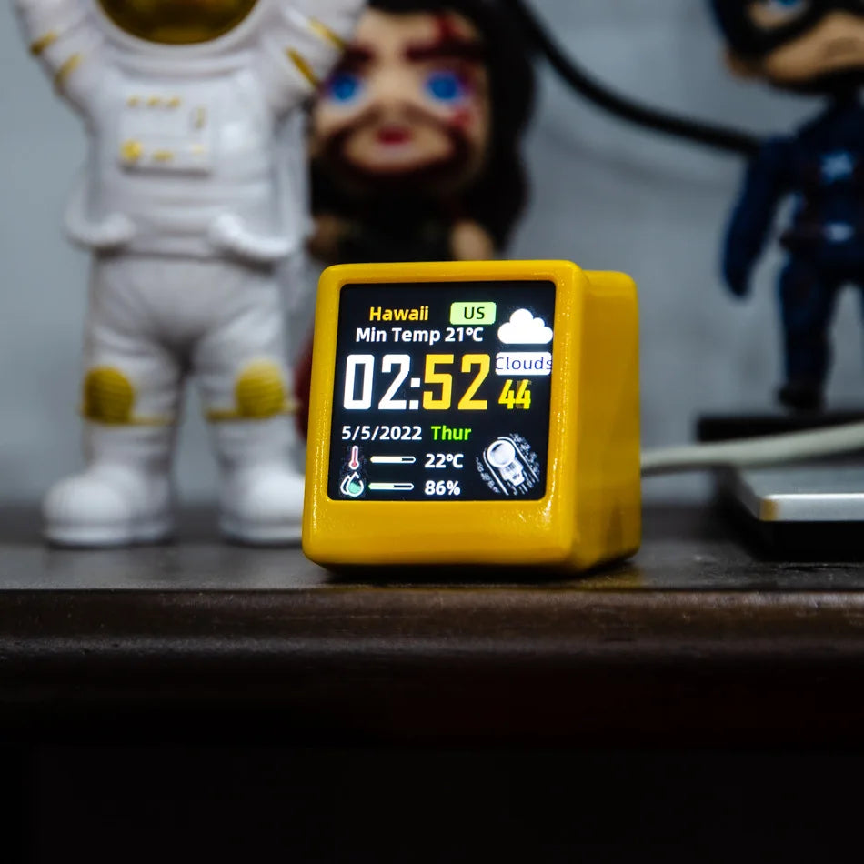 Digital clock with weather display on a wooden surface, with figurines in the background.