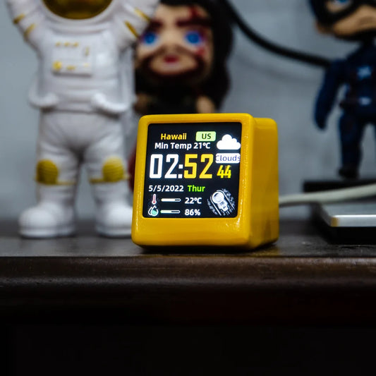 Digital clock with weather display on a wooden surface, with figurines in the background.