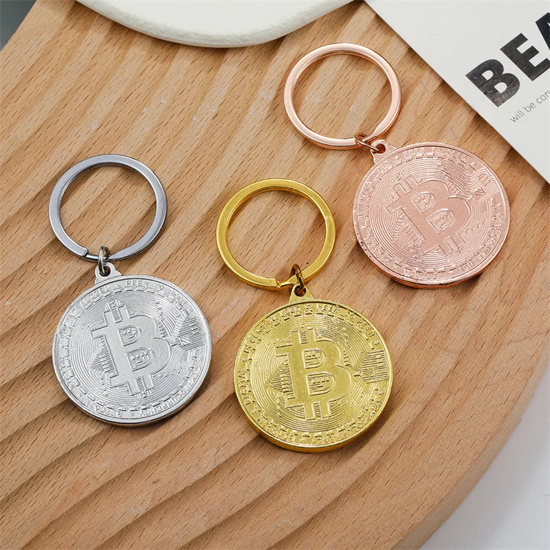 Three Bitcoin-themed keychains in silver, gold, and rose gold on a wooden surface.