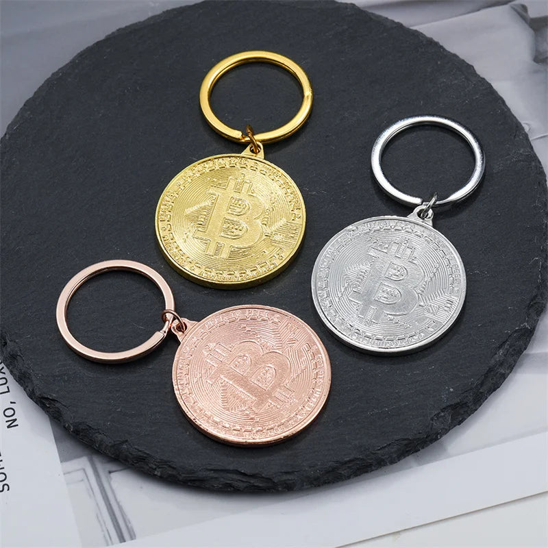 Three Bitcoin-themed keychains in gold, silver, and rose gold on a dark slate surface.