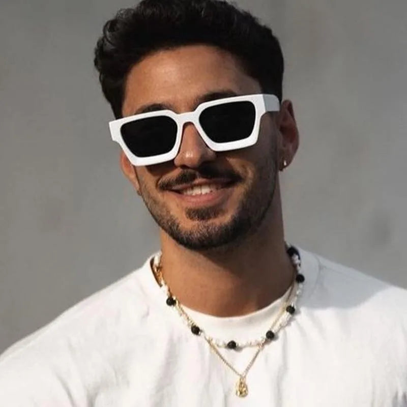 Man wearing white sunglasses and a white shirt with layered necklaces against a gray background