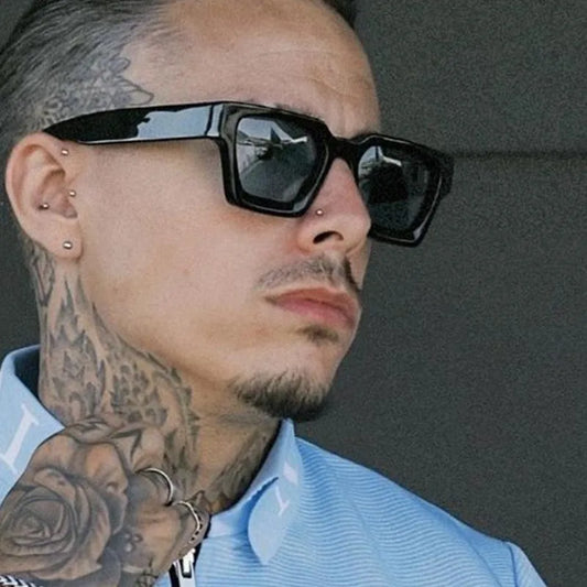 Man with super stylish block sunglasses dark shades, tattoos and a blue shirt against a gray background.