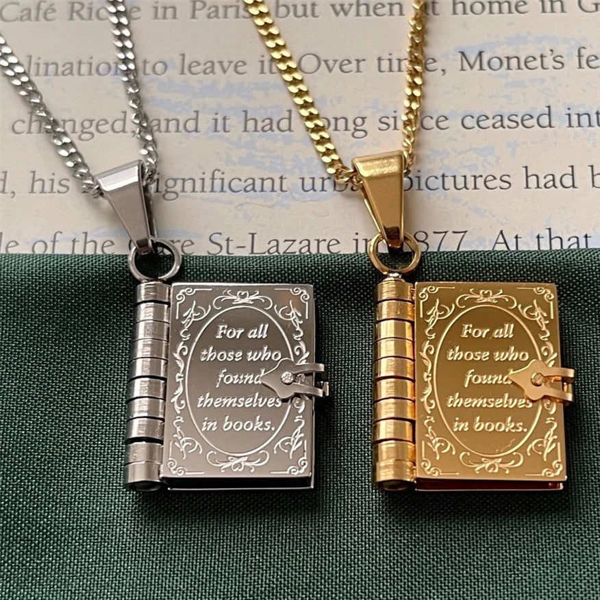 Two book-shaped pendant necklaces on chains, one silver-toned and one gold-toned, both engraved with the quote 'For all those who found themselves in books.' They are displayed side by side on a dark green textured surface with a book page in the background.