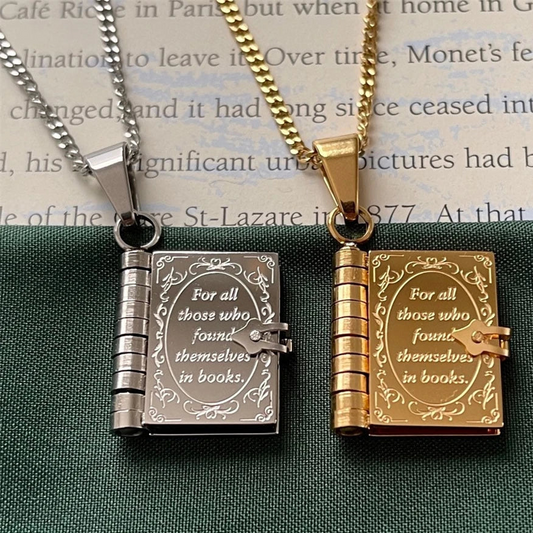 Two book-shaped pendant necklaces on chains, one silver-toned and one gold-toned, both engraved with the quote 'For all those who found themselves in books.' They are displayed side by side on a dark green textured surface with a book page in the background.