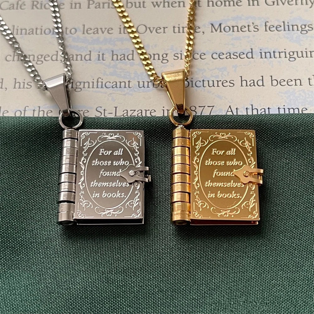Two book-shaped pendants, one silver and one gold, on a textured surface with text.