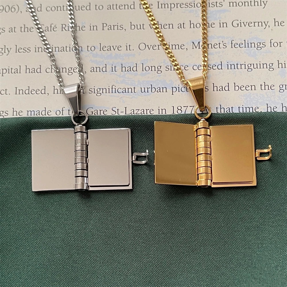 Two book-shaped pendants, one silver and one gold, on a green surface with text in the background.