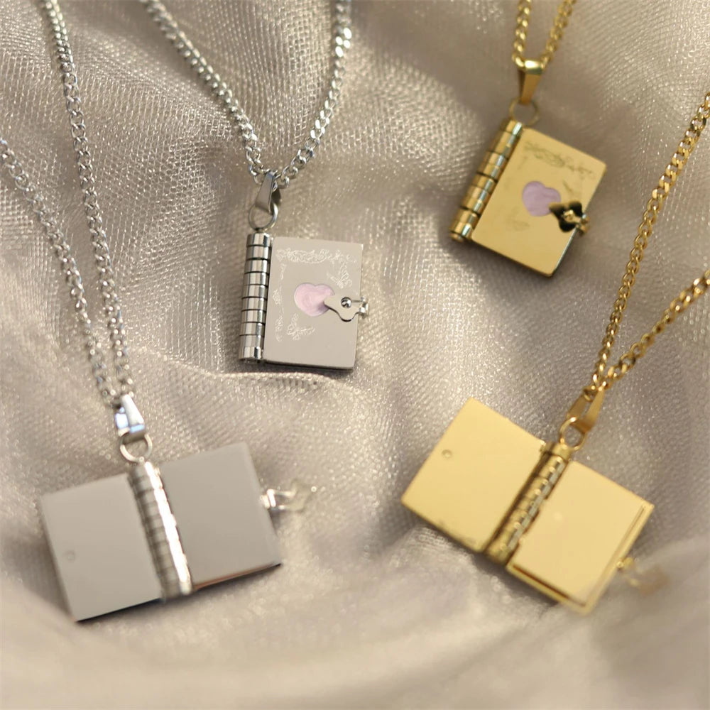 Four book-shaped pendants on necklaces, in silver and gold, on a soft fabric background.
