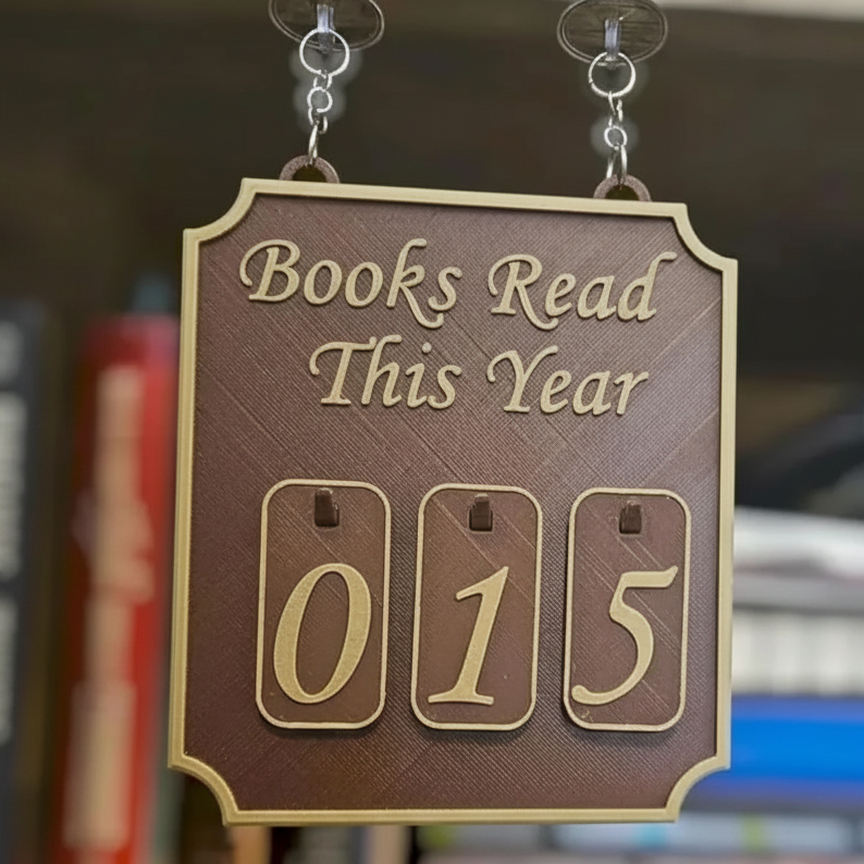A decorative hanging plaque with the phrase “Books Read This Year” and three flip-counting numerals showing “015,” suspended by clear clips against a blurred bookshelf background.