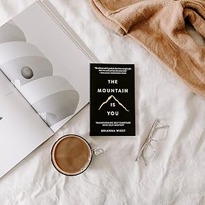 Book titled 'The Mountain is You' by Brianna Wiest on a white surface with a cup of coffee and a blanket.