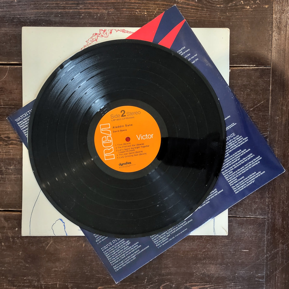 A black vinyl record with a bright orange center label reading RCA Victor, placed on top of printed paper sleeves on a dark wood surface.
