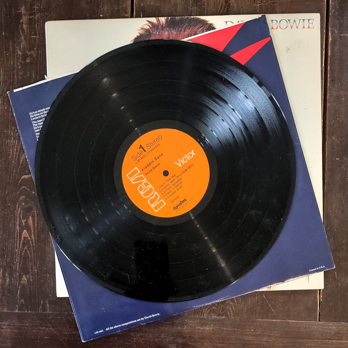 Black vinyl record of Aladdin Sane with an orange Victor label lying on top of a blue and cream David Bowie album cover, photographed on a dark wood surface.