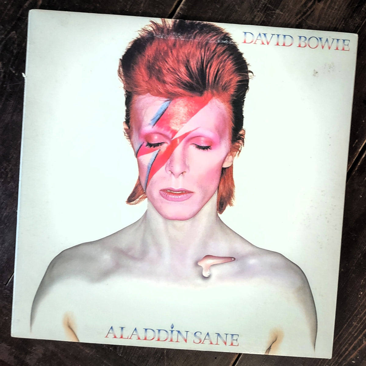 David Bowie 'Aladdin Sane' vinyl record album cover, photographed flat on a dark wooden surface. The cover features David Bowie with his signature lightning-reflector makeup, and the text 'DAVID BOWIE' and 'ALADDIN SANE' is printed on the cover.