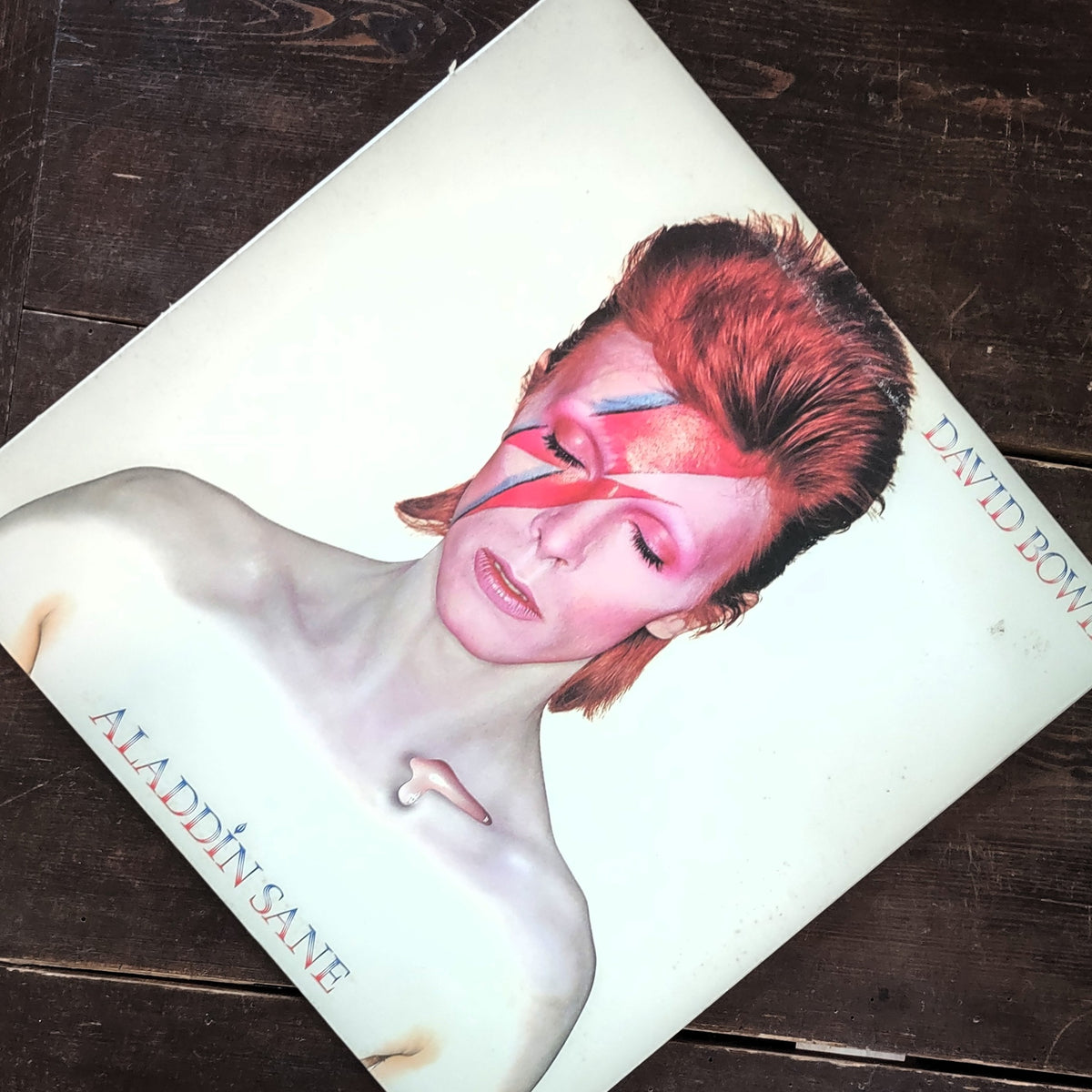 David Bowie's 'Aladdin Sane' vinyl record sleeve photographed diagonally on a dark wooden surface. The cover features a portrait of Bowie with pink and blue lightning-embroidered makeup, with the title 'ALADDIN SANE' and 'DAVID BOWIE' printed in blue text.