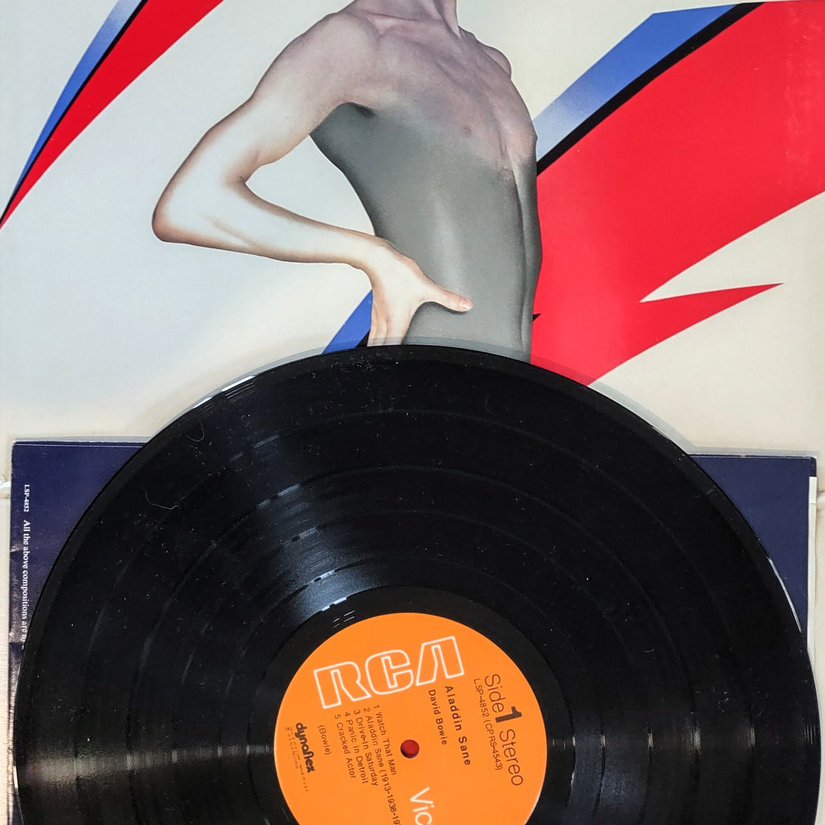 David Bowie Aladdin Sane vinyl record partially pulled from its sleeve, with the black record dominating the foreground and the sleeve artwork behind it showing a stylized nude torso against red, white, and blue graphic shapes. The center label reads RCA Side-1.