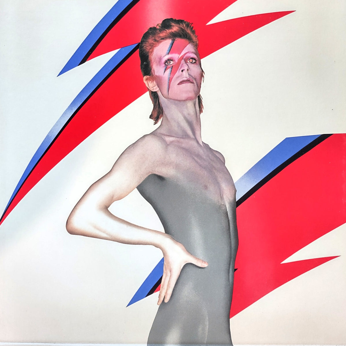 David Bowie in a metallic silver body suit with red and blue lightning-arc face paint, posed against a white background with large red and blue lightning-bolt graphics.