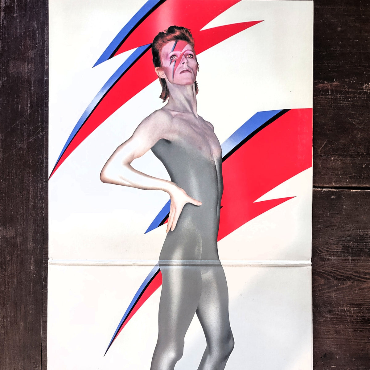 A vintage Aladdin Sane album insert featuring David Bowie in a metallic silver bodysuit against a white background with bold red and blue lightning bolt graphics, displayed on a dark wooden surface.