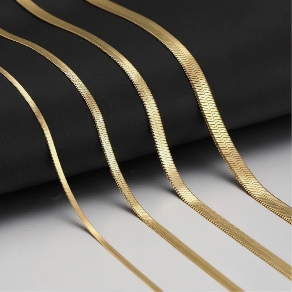 Four gold snake herringbone chain necklaces of varying thicknesses draped elegantly over a black textured background, with a white surface visible in the lower right corner.