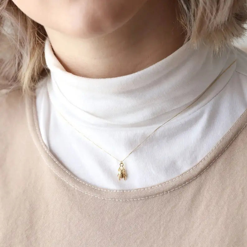 Gold scarab beetle necklace with a small pendant worn by a person in a white turtleneck and beige cardigan.